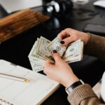 Unrecognizable elegant female in sweater counting dollar bills while sitting at wooden table with planner and pen